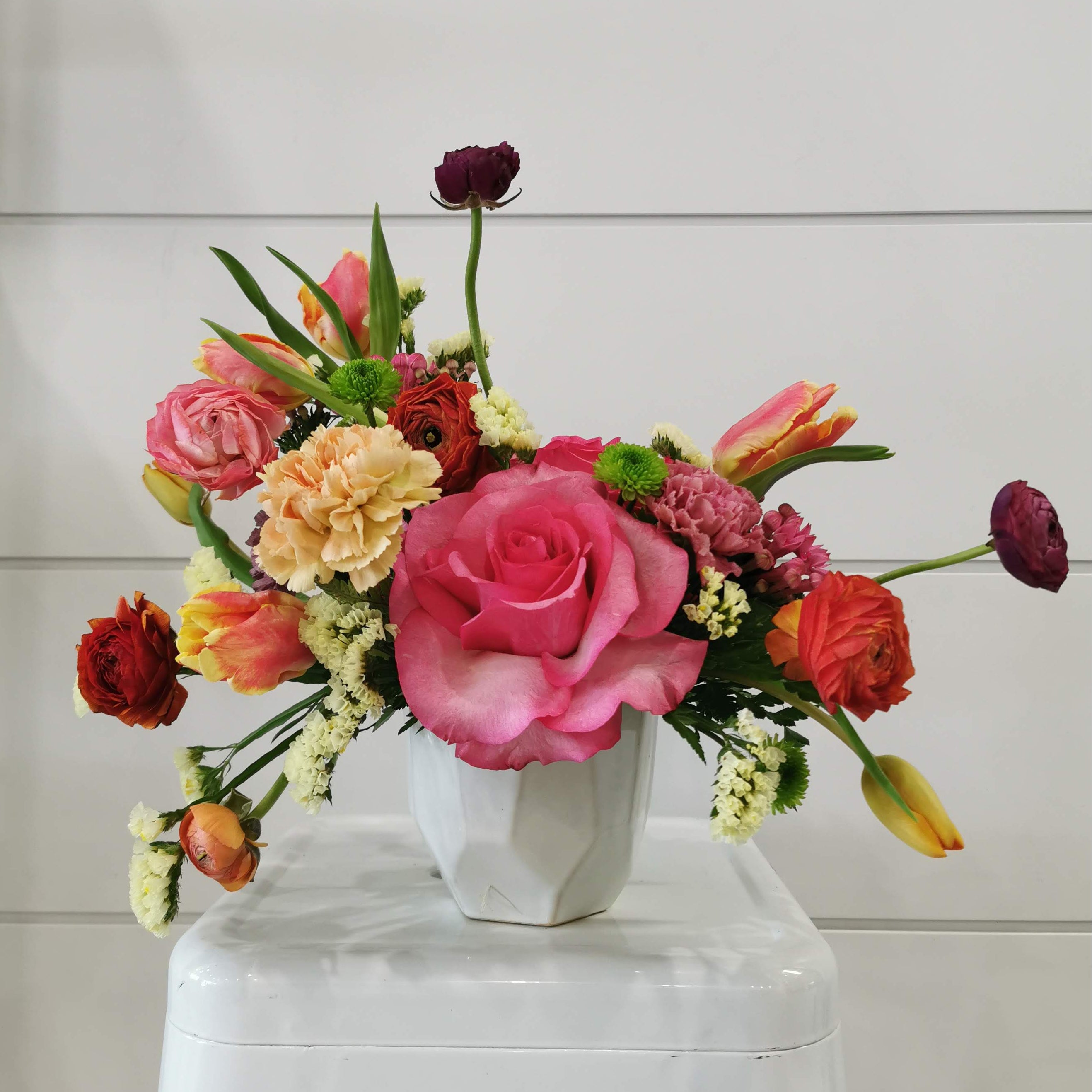 A vibrant floral arrangement featuring a large pink rose, red and orange tulips, yellow peonies, and green foliage, presented in a white geometric ceramic vase on a white stool against a white paneled wall.