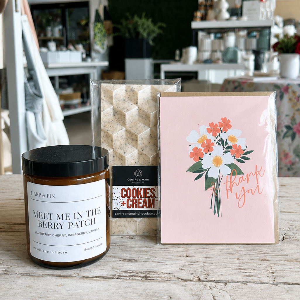 Candle, cookies & cream package, and card on a wooden surface with a store in the background.