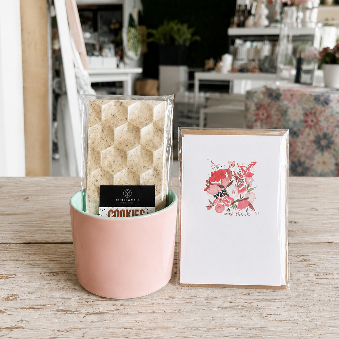 Tri colour mug with decorative stones, a chocolate bar and a floral card on a wooden surface.